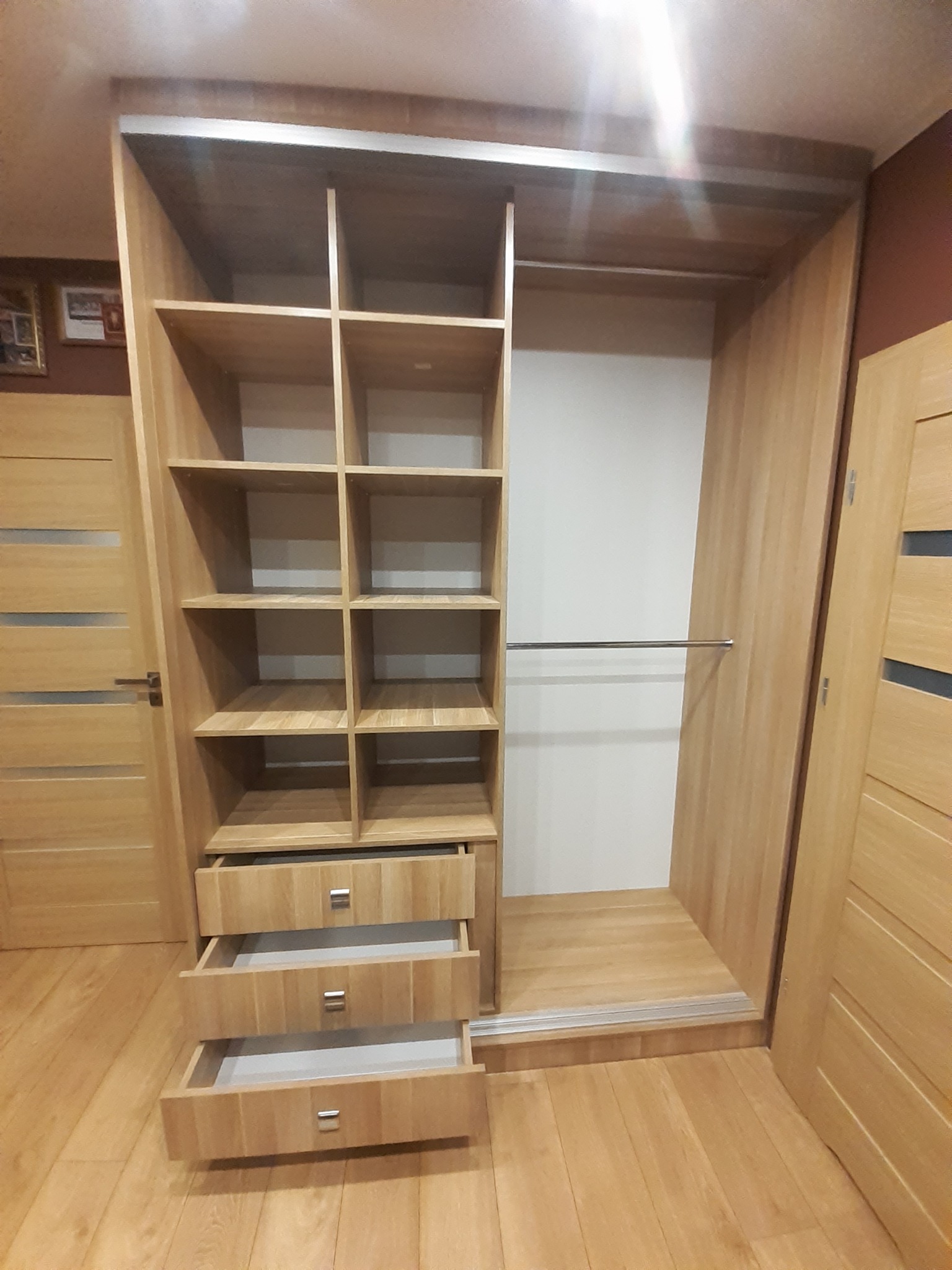 Jasna, drewniana szafa wnękowa z otwartymi półkami i wysuwanymi szufladami, widoczna w całości, z metalowymi uchwytami, komponująca się z podłogą i drzwiami.