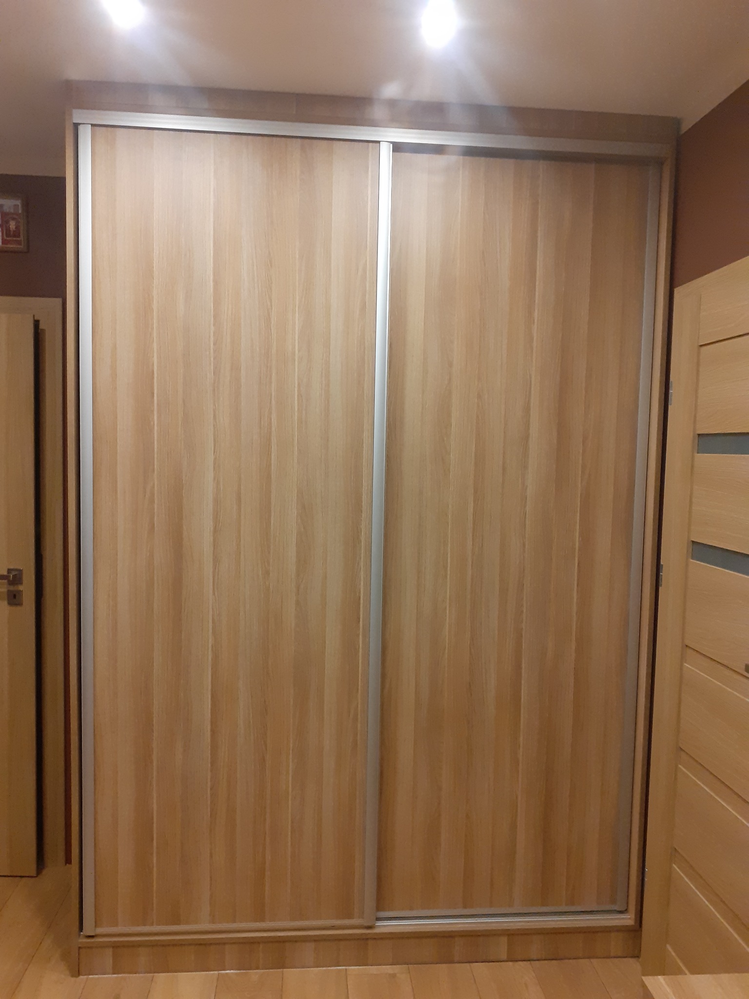 Jasna, dwudrzwiowa szafa przesuwna z drewnopodobnymi frontami i srebrnymi profilami, dopasowana do wnętrza z drewnianą podłogą i drzwiami.