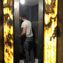 BUMAR Mariusz Ambroży - Pracownik czyści ściany wyłożone płytami marmuru w kabinie prysznicowej z podświetlanym onyksem w witrynie, widoczna surowa żarówka i niedokończone fugowanie.