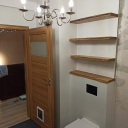 BUMAR Mariusz Ambroży - Łazienka w trakcie wykańczania: białe płytki, drewniane półki o nieregularnych krawędziach, wisząca toaleta z czarnym przyciskiem, chromowany żyrandol i drewniane drzwi z szybą i otworem dla zwierząt.