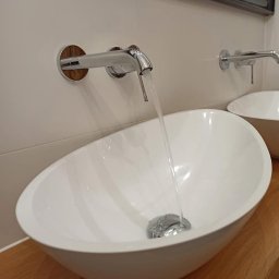 BUMAR Mariusz Ambroży - Biała, owalna umywalka nablatowa z chromowaną armaturą ścienną z drewnianymi rozetami, z której leje się woda, ustawiona na drewnianym blacie.