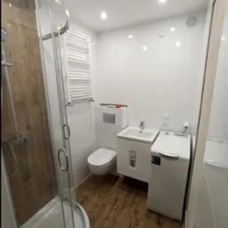 Niewielka, jasna łazienka w bloku z kabiną prysznicową z drewnopodobnymi panelami, białą umywalką z szafką, podwieszaną toaletą, grzejnikiem drabinkowym i pralką.