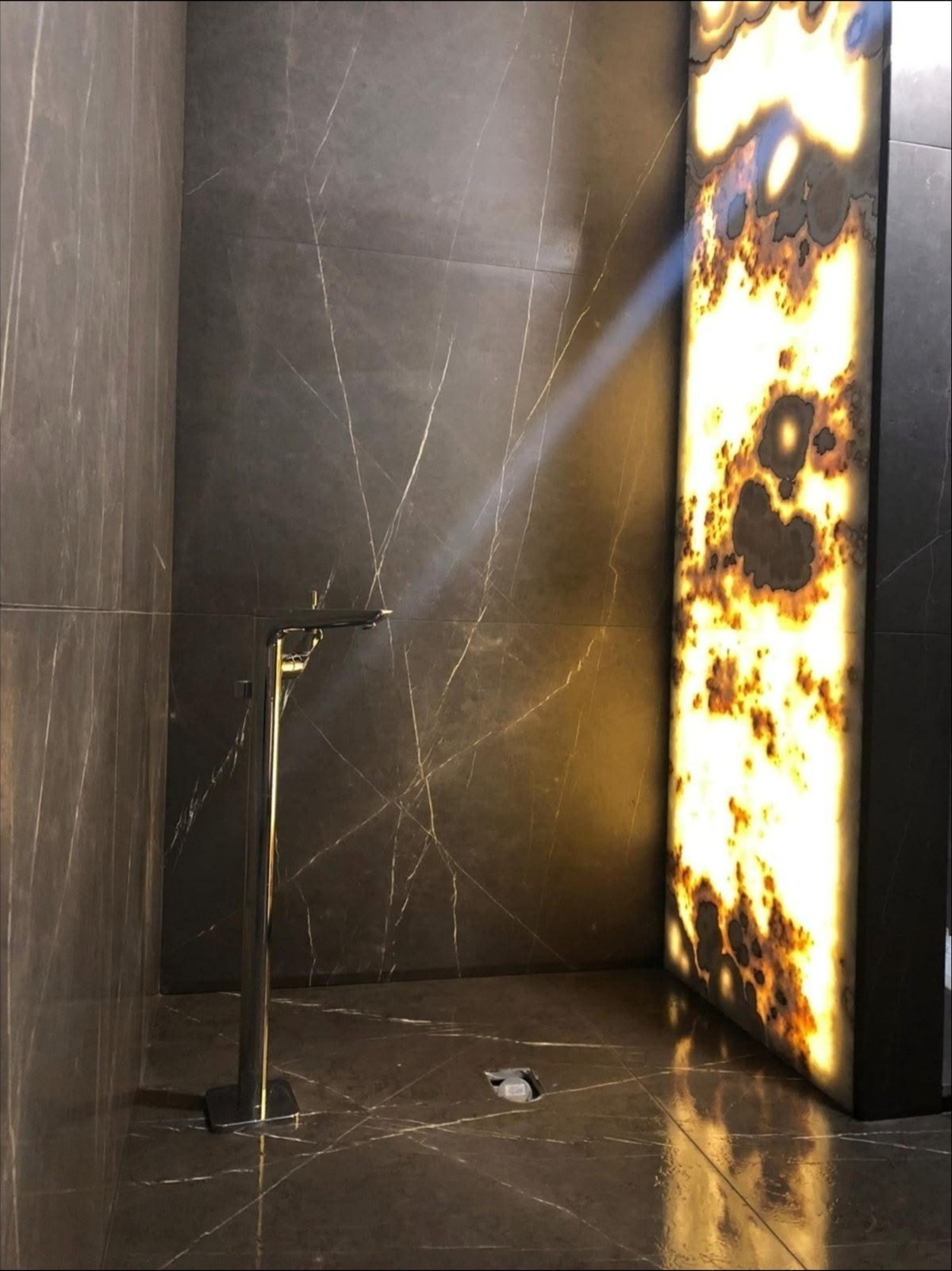 Nowoczesna łazienka wykończona włoskim marmurem w płytach, z podświetlonym panelem z onyksu i minimalistyczną, chromowaną baterią podłogową.