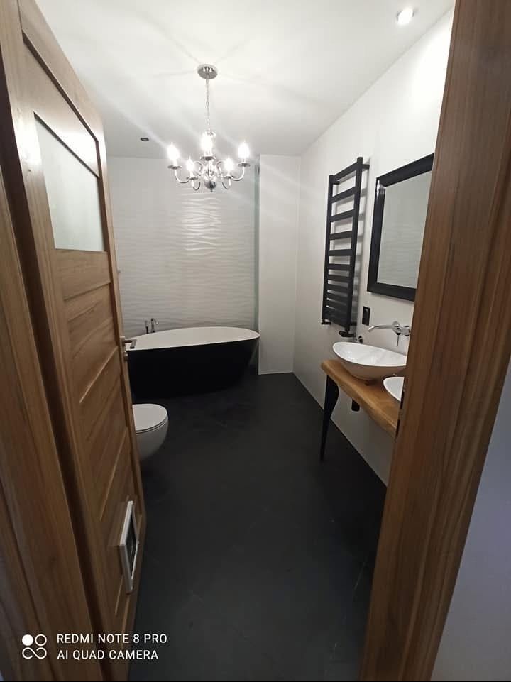 Nowoczesna łazienka z czarną podłogą, wolnostojącą wanną i drewnianą umywalką, widok zza drewnianych drzwi, oświetlona przez dekoracyjny żyrandol.