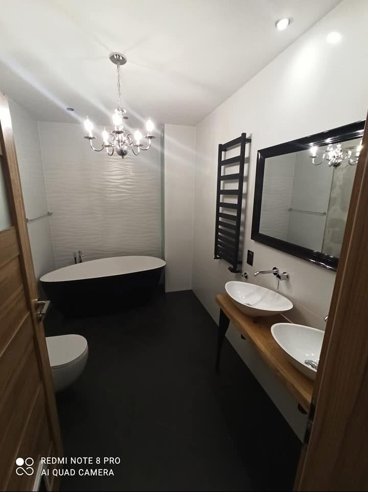Nowoczesna łazienka z wolnostojącą czarno-białą wanną, dwoma umywalkami na drewnianym blacie z czarnymi nogami, czarnym grzejnikiem drabinkowym i lustrem w czarnej ramie, oświetlona żyrandolem.