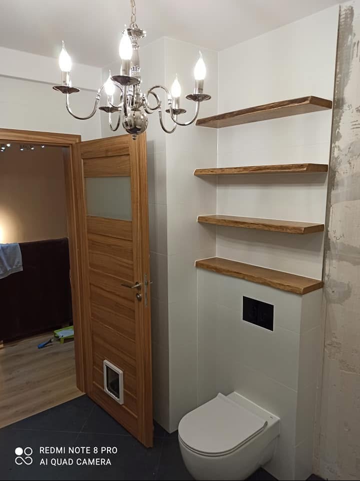 Łazienka w trakcie wykańczania: białe płytki, drewniane półki o nieregularnych krawędziach, wisząca toaleta z czarnym przyciskiem, chromowany żyrandol i drewniane drzwi z szybą i otworem dla zwierząt.