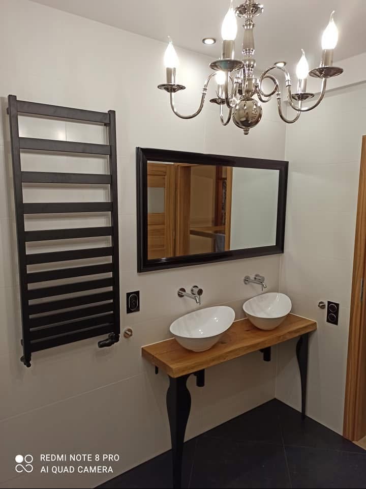 Nowoczesna łazienka z dwoma białymi umywalkami nablatowymi na drewnianym blacie z czarnymi nogami, czarny grzejnik drabinkowy na ścianie, bateria umywalkowa ścienna, czarne płytki podłogowe, lustro...