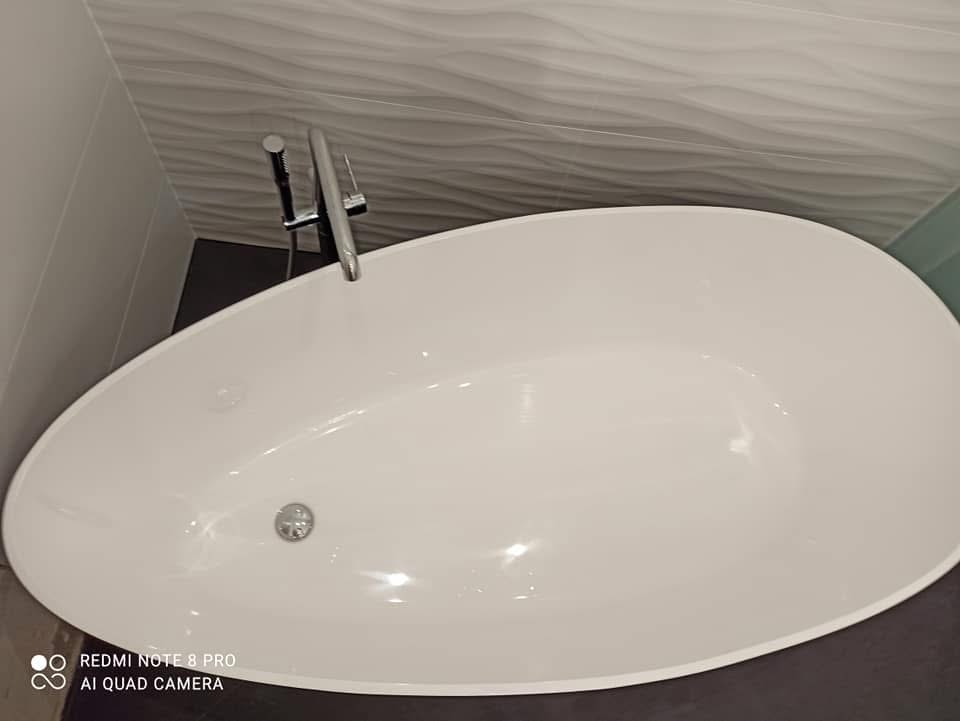 Biała wanna wolnostojąca z chromowaną baterią podtynkową, na tle ściany wykończonej płytkami o falującej strukturze.