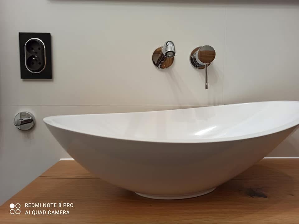 Biała, owalna umywalka nablatowa na drewnianym blacie, z chromowaną armaturą montowaną w ścianie i gniazdkiem elektrycznym w czarnej ramce.