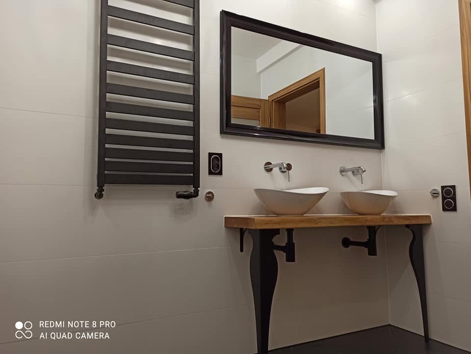 Nowoczesna łazienka z podwójną umywalką w kształcie misy na blacie z surowego drewna wspartym na czarnych nogach, baterie umywalkowe ścienne, grzejnik drabinkowy w kolorze grafitowym, lustro...