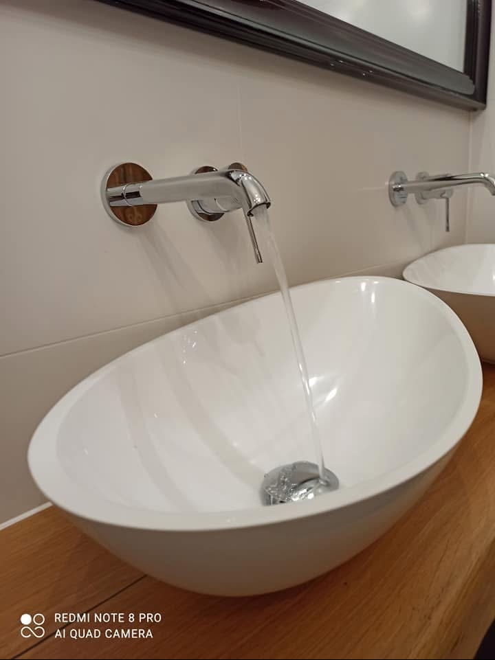 Biała, owalna umywalka nablatowa z chromowaną armaturą ścienną z drewnianymi rozetami, z której leje się woda, ustawiona na drewnianym blacie.