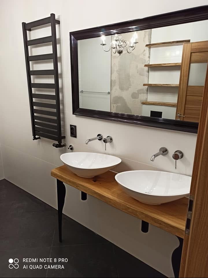 Nowoczesna łazienka z dwoma białymi umywalkami nablatowymi na blacie z surowego drewna, czarne nogi blatu, baterie ścienne, duże lustro w ciemnej ramie odbijające żyrandol i półki, grzejnik...