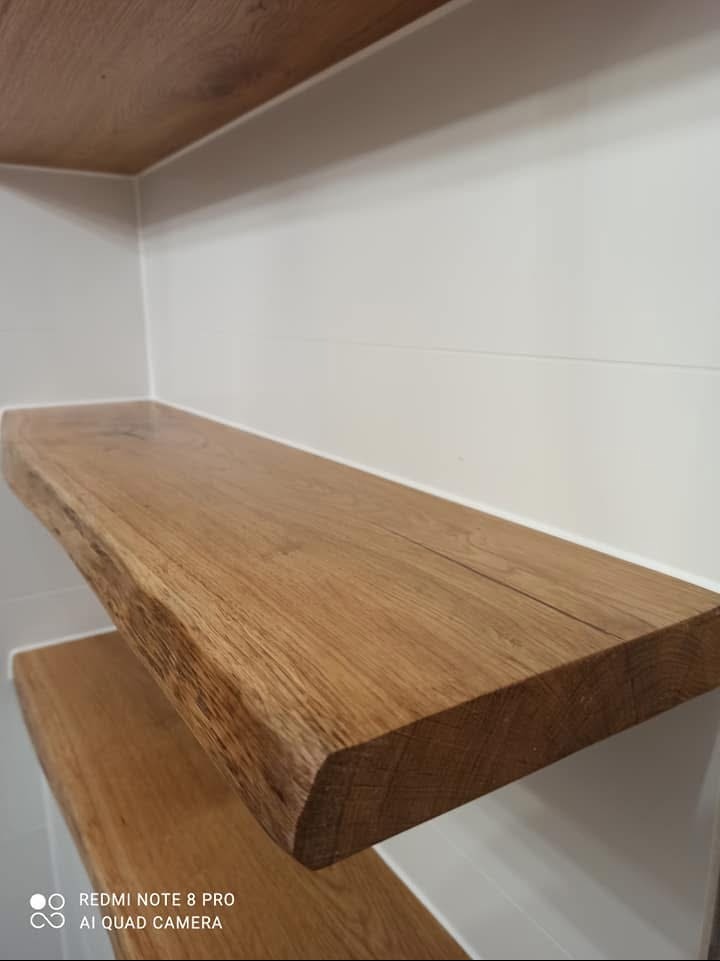 Dwie półki z surowego drewna zamontowane na białej ścianie wyłożonej płytkami w łazience. Krawędzie półek nieregularne, zachowujące naturalny wygląd drewna.