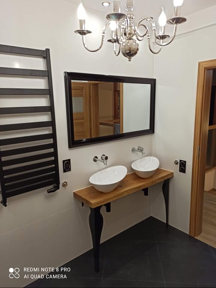 Nowoczesna łazienka z dwoma okrągłymi umywalkami nablatowymi na blacie z surowego drewna, czarne, gięte nogi, bateria umywalkowa ścienna, czarny grzejnik drabinkowy, lustro w czarnej ramie...