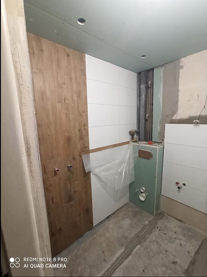 Wnętrze łazienki w trakcie prac wykończeniowych: Częściowo ułożone płytki imitujące drewno, białe płytki ścienne, zabudowa podtynkowego WC z płyt gipsowo-kartonowych, odkryte rury instalacyjne.