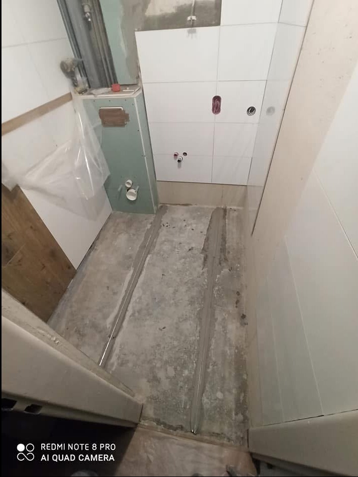 Wnętrze łazienki w trakcie prac wykończeniowych: widoczne białe płytki na ścianie, przygotowana instalacja wodno-kanalizacyjna, surowa podłoga z wyznaczonymi liniami, zabudowa geberitu w trakcie...