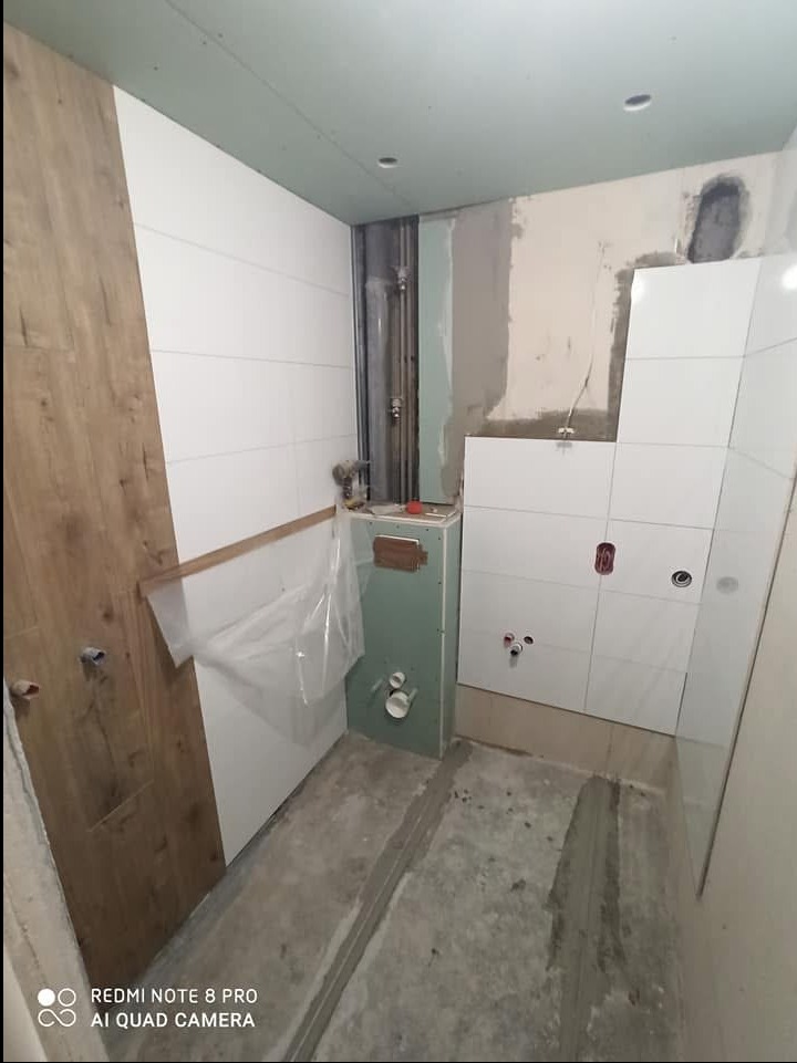 Wnętrze łazienki w trakcie prac wykończeniowych: częściowo ułożone białe płytki ścienne, fragment ściany z okładziną drewnopodobną, widoczne rury i instalacje, surowy beton na podłodze.