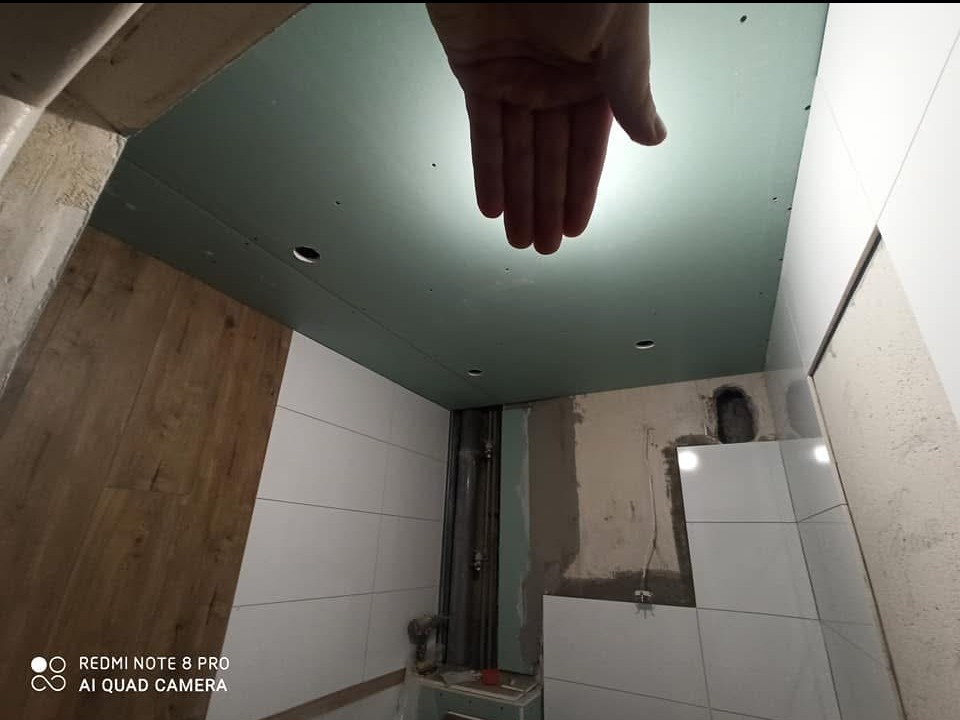 Remont łazienki w trakcie: widoczny sufit z płyt gipsowo-kartonowych z wyciętymi otworami na oświetlenie, częściowo wykończona ściana z białymi płytkami oraz fragment ściany z okładziną...