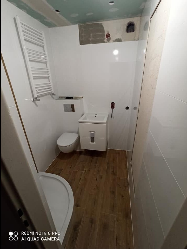 Mała łazienka w trakcie wykańczania, widoczne białe płytki na ścianach, drewnopodobne płytki na podłodze, biała umywalka z szafką, toaleta, grzejnik drabinkowy, sufit częściowo wykończony płytami...