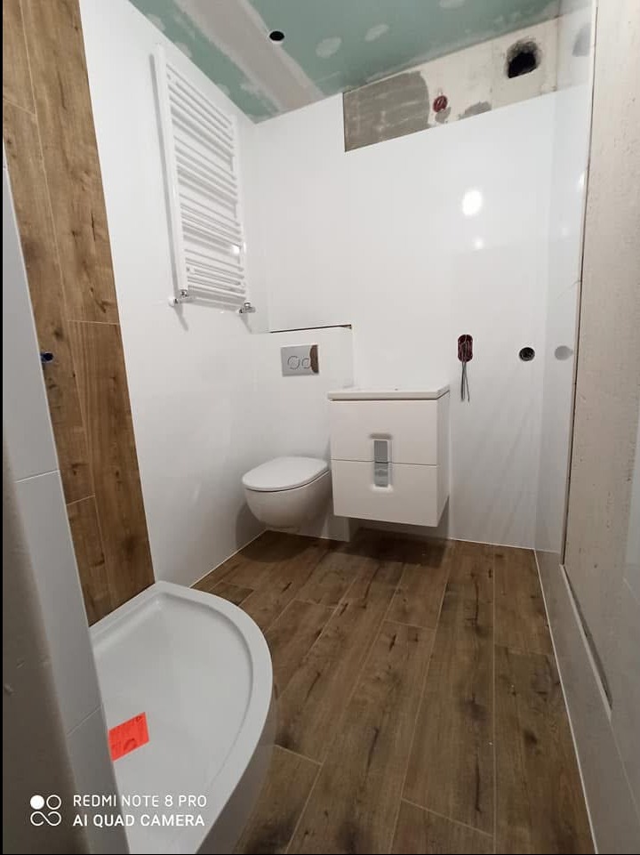 Nieduża łazienka w trakcie prac wykończeniowych: białe ściany z fragmentami surowego tynku, toaleta podwieszana, szafka umywalkowa, grzejnik drabinkowy i kabina prysznicowa z brodzikiem, płytki...