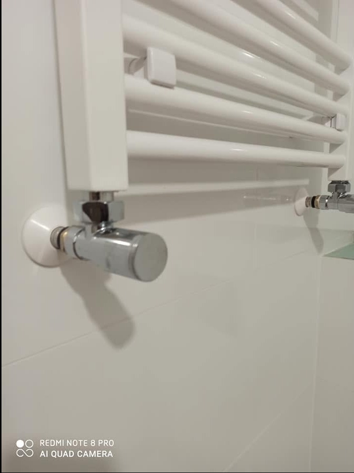 Biały grzejnik łazienkowy drabinkowy zamontowany na białych, błyszczących płytkach, widoczne chromowane zawory i przyłącza.