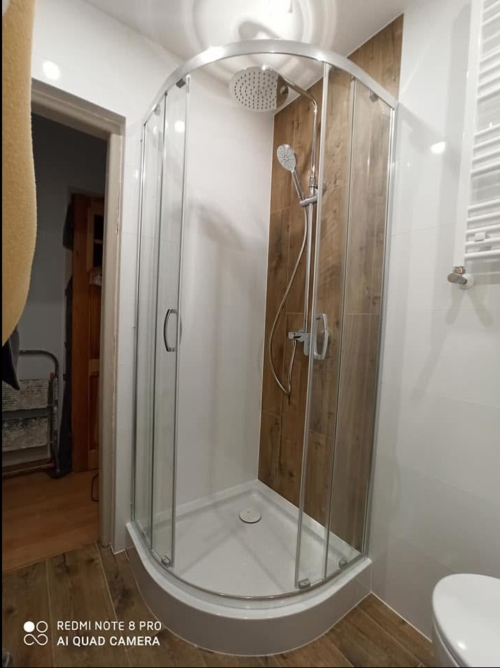 Narożna kabina prysznicowa z przesuwnymi drzwiami, wykończona płytkami drewnopodobnymi na jednej ze ścian i białymi płytkami na pozostałych, z widocznym grzejnikiem drabinkowym i podłogą imitującą...