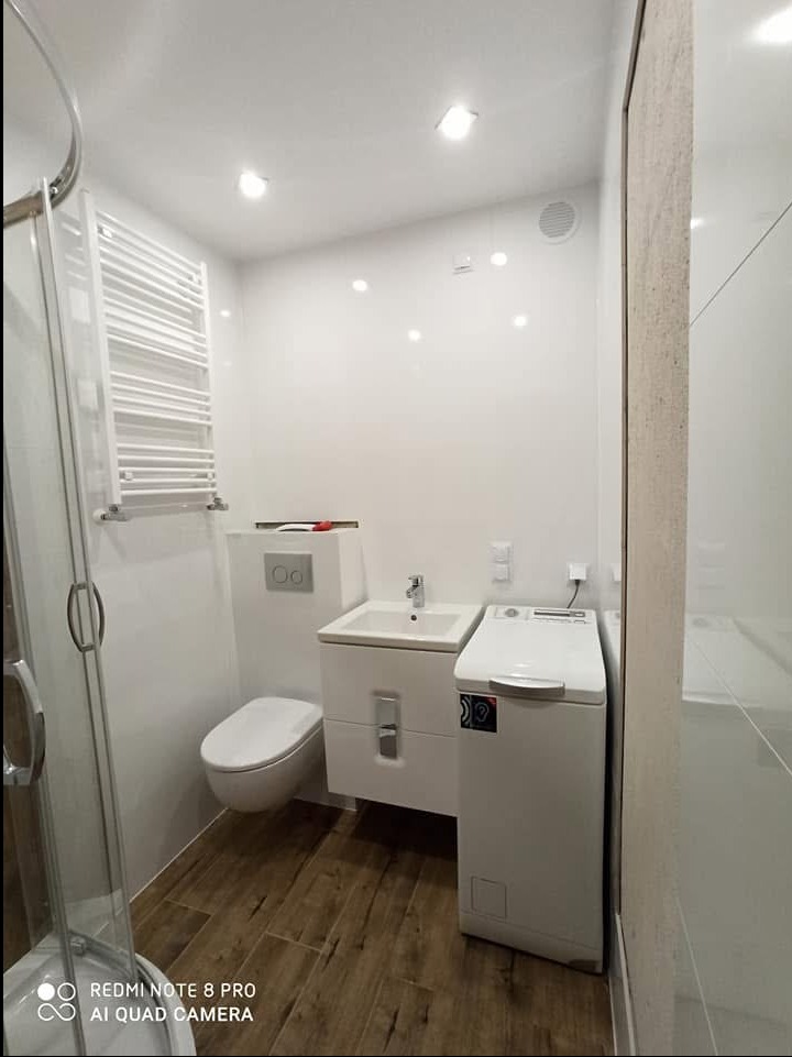 Niewielka, ale funkcjonalna łazienka w bloku z białymi płytkami ściennymi i podłogą imitującą drewno, wyposażona w kabinę prysznicową, umywalkę z szafką, toaletę podwieszaną i pralkę.