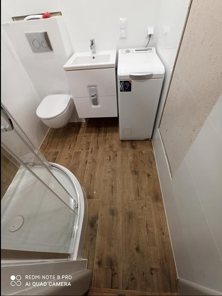 Niewielka łazienka z kabiną prysznicową, podwieszaną toaletą, umywalką z szafką i pralką. Na podłodze płytki imitujące drewno, ściany białe.