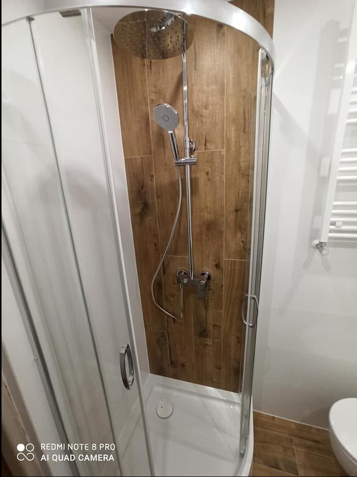 Narożna kabina prysznicowa z transparentnymi drzwiami na tle ściany wyłożonej płytkami drewnopodobnymi, bateria natryskowa z deszczownicą i słuchawką, oraz biały grzejnik drabinkowy w małej łazience.