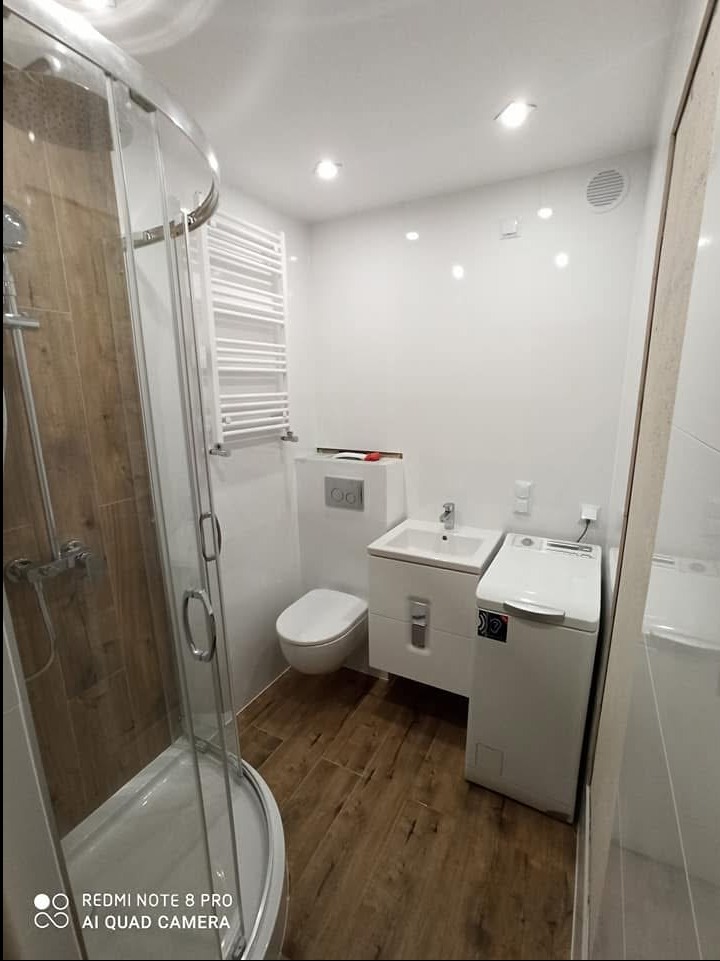 Niewielka, jasna łazienka w bloku z kabiną prysznicową z drewnopodobnymi panelami, białą umywalką z szafką, podwieszaną toaletą, grzejnikiem drabinkowym i pralką.