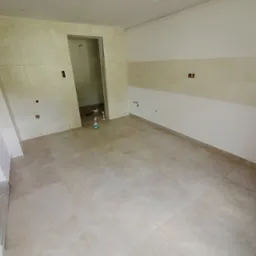 Wykończona łazienka z beżowymi płytkami na podłodze i ścianach do połowy wysokości. Widoczne puszki elektryczne i surowa żarówka. W głębi pomieszczenia otwarte przejście.