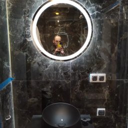 Dawid Czernik DAVMONT - Czarna umywalka nablatowa i okrągłe lustro LED na tle marmurowych płytek. Widoczne gniazdka elektryczne. W lustrze odbicie osoby robiącej zdjęcie.