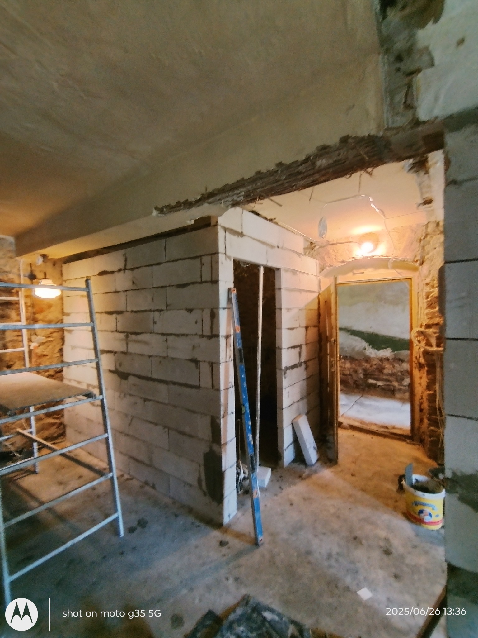 Wnętrze w trakcie remontu: ściana z białych pustaków, poziomica oparta o ścianę, drabina, surowe wykończenie, widoczne instalacje. Prace murarskie w toku.