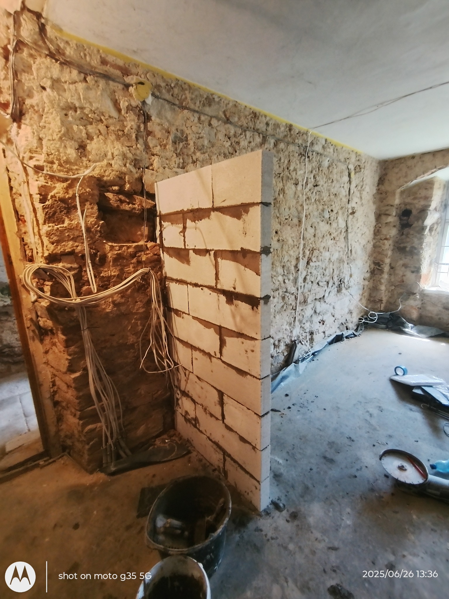 Wnętrze w trakcie renowacji: surowe kamienne ściany, nowa ścianka działowa z bloczków i zwisające przewody elektryczne. Widoczne narzędzia i wiadro.