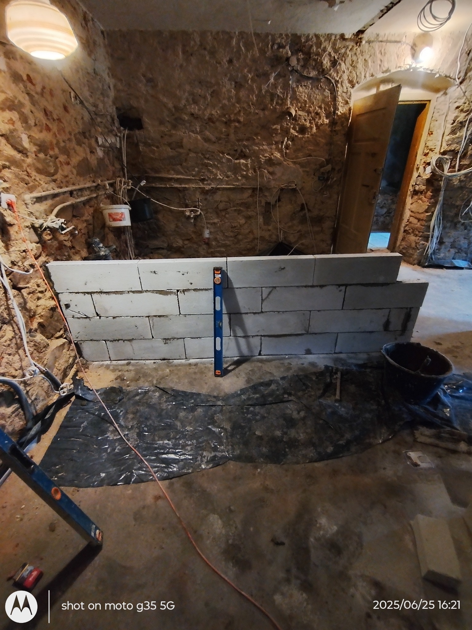 Murowanie ściany z bloczków w surowym wnętrzu, widoczny poziomica i wiadro, prace w toku. Kamienne ściany w tle.