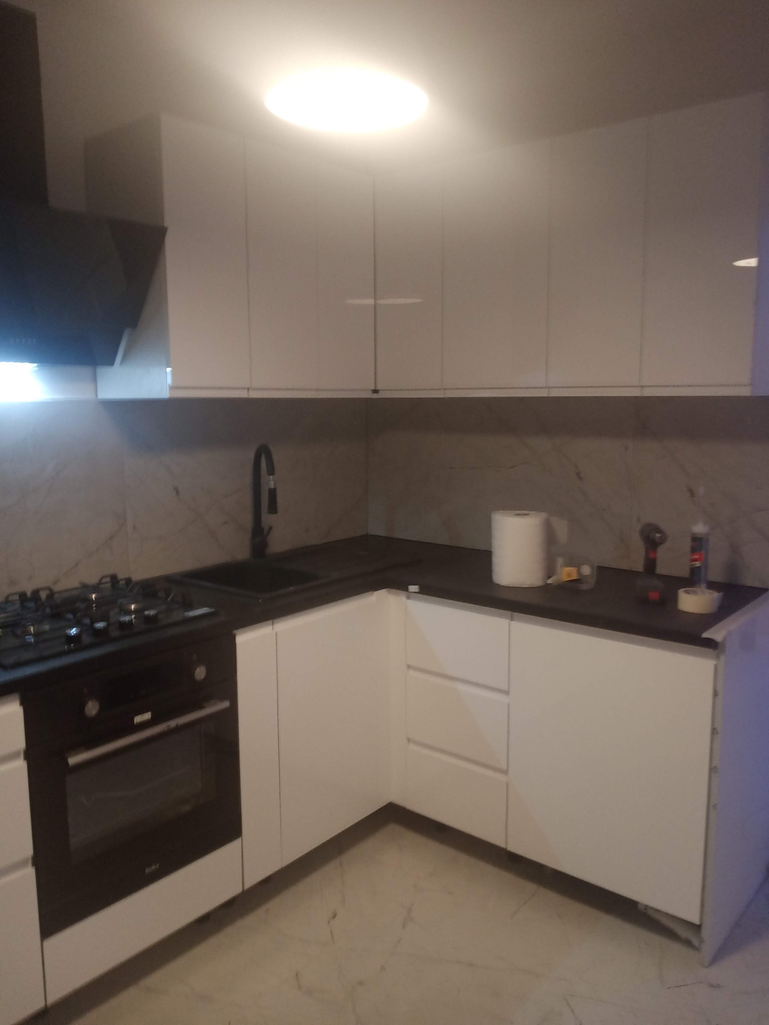 Nowoczesna kuchnia z białymi szafkami, czarnym blatem i płytą gazową. Widoczna bateria i narzędzia na blacie. Marmurowe wykończenie ściany nad blatem.