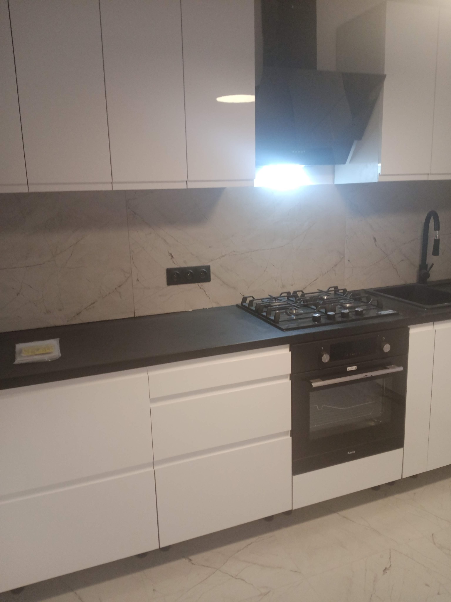 Nowoczesna kuchnia z czarnym blatem i białymi szafkami w Nowej Rudzie. Widoczna płyta gazowa, piekarnik Amica i marmurowy backsplash.