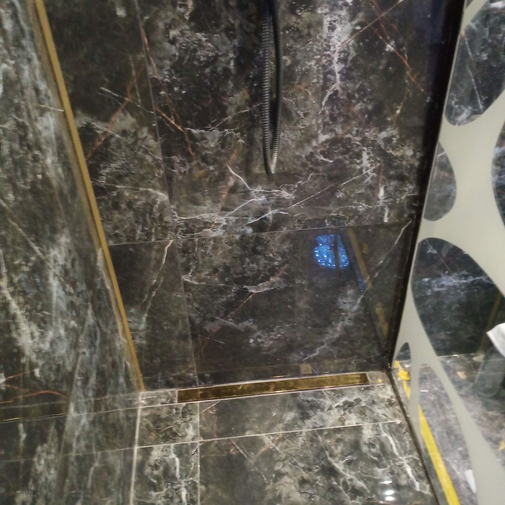 Czarna, marmurowa kabina prysznicowa z odpływem liniowym w złotym kolorze i szklaną ścianką z dekoracyjnymi wycięciami w kształcie kół.