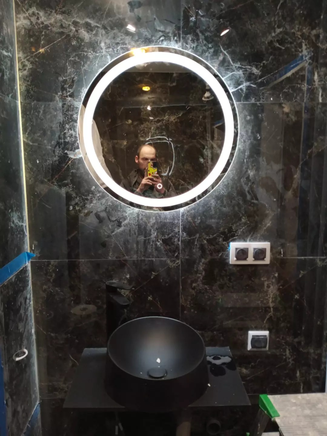 Czarna umywalka nablatowa i okrągłe lustro LED na tle marmurowych płytek. Widoczne gniazdka elektryczne. W lustrze odbicie osoby robiącej zdjęcie.