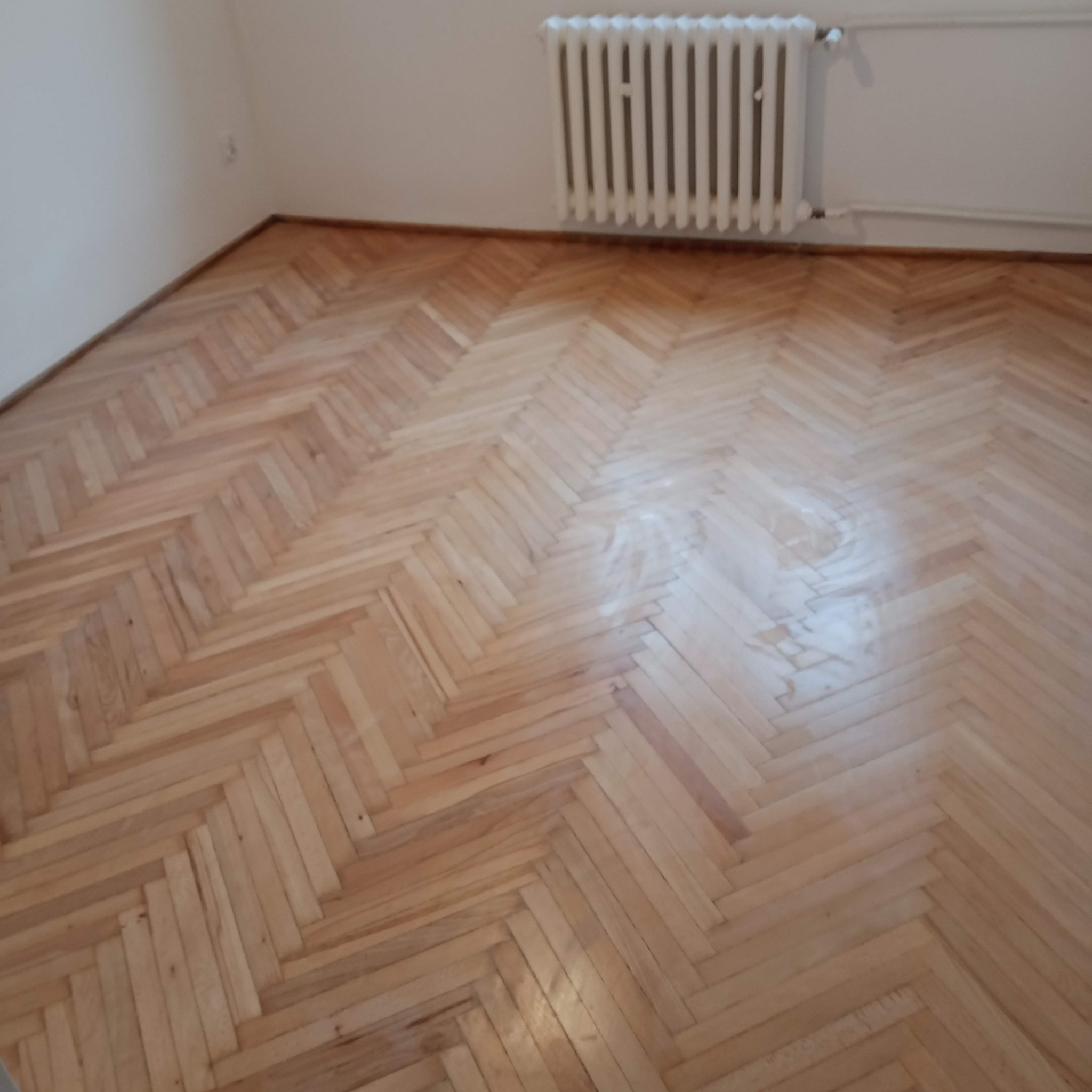 Ukończona podłoga z parkietu ułożonego w jodełkę, widoczny grzejnik na ścianie. Jasne drewno i proste ściany tworzą minimalistyczny, wykończony wygląd wnętrza.