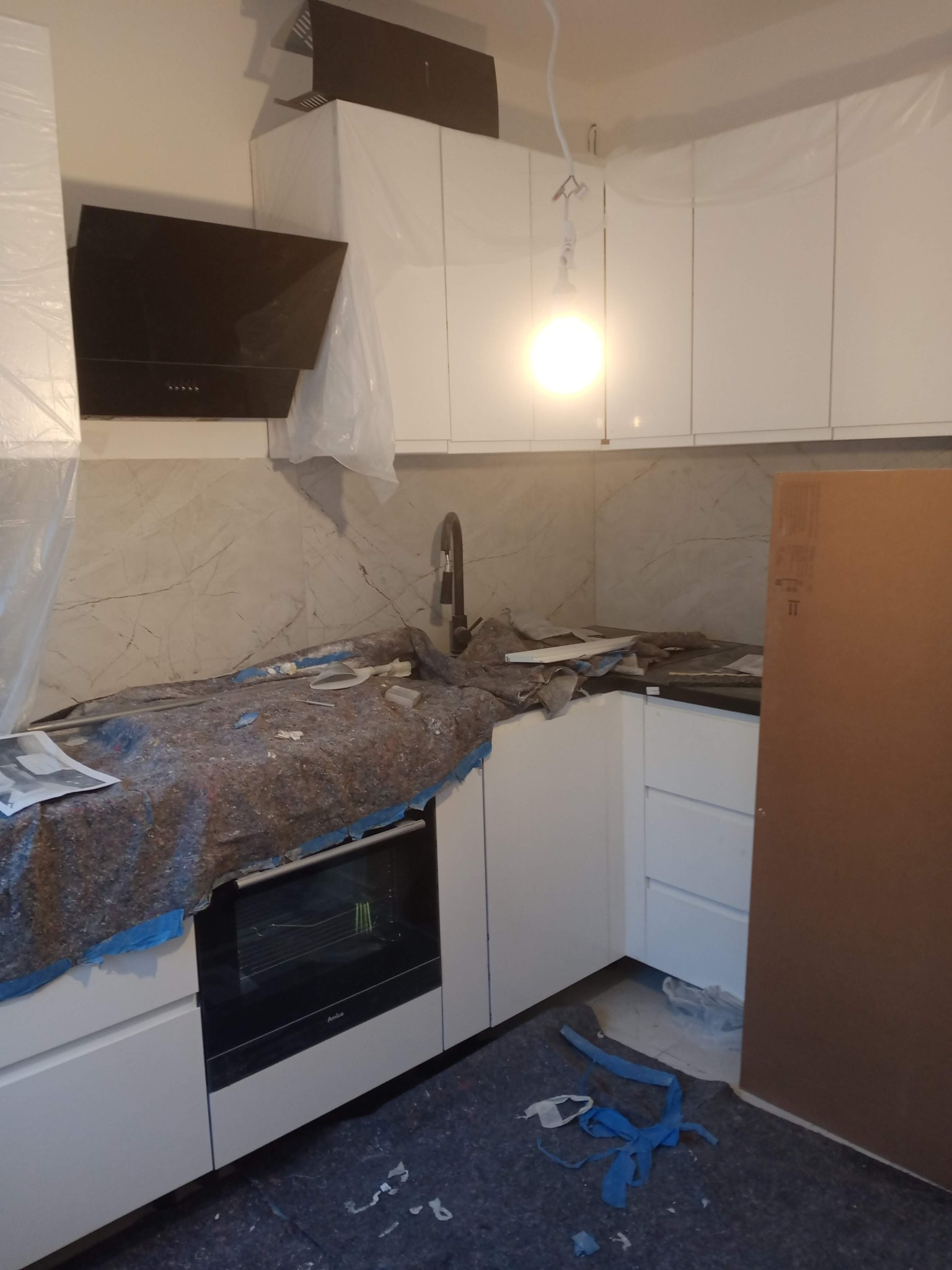 Kuchnia w trakcie montażu: białe szafki, czarny okap, marmurowe ściany, zabezpieczone blaty i podłoga. Widoczny fragment piekarnika Amica.