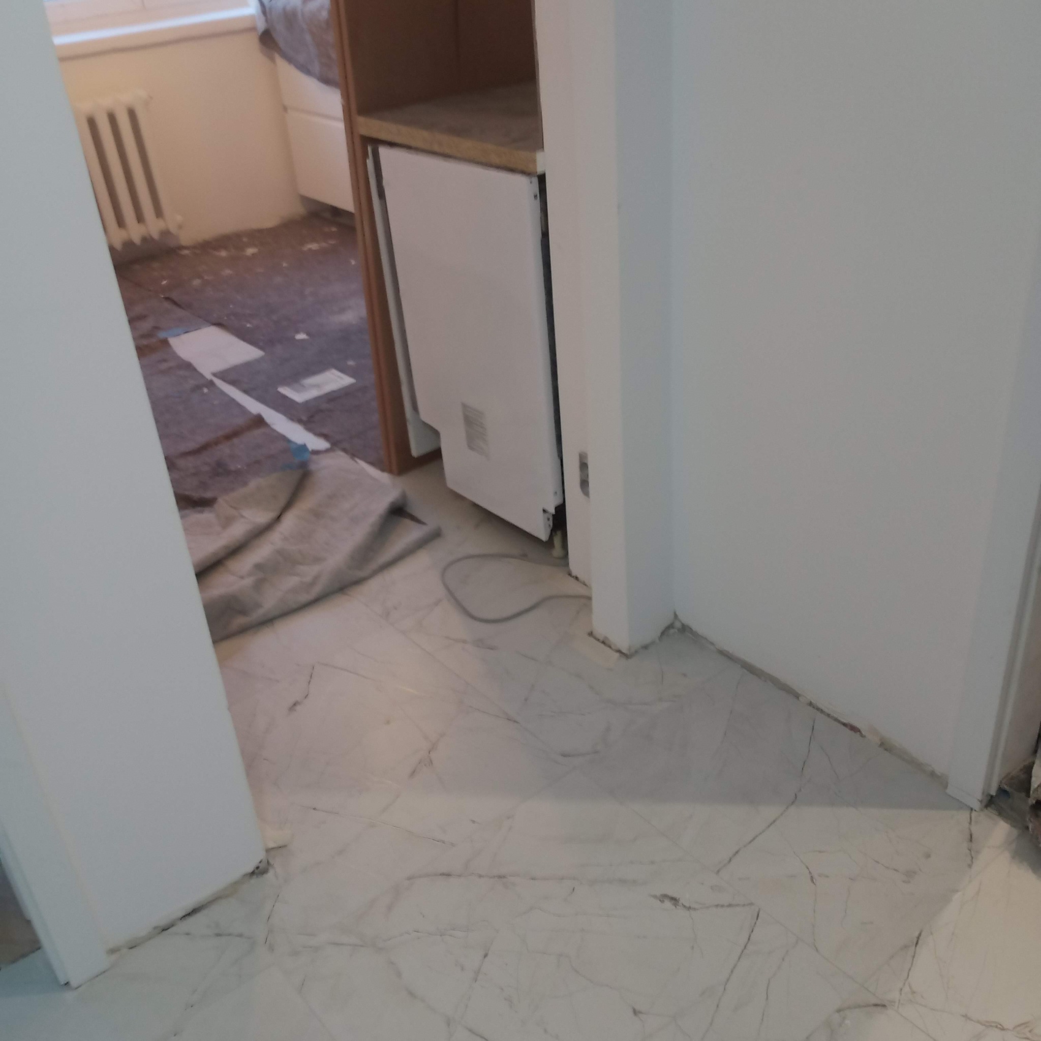 Biała marmurowa podłoga w trakcie instalacji, widoczny fragment grzejnika i szafki z niedokończonymi pracami wykończeniowymi. Kabel na podłodze.