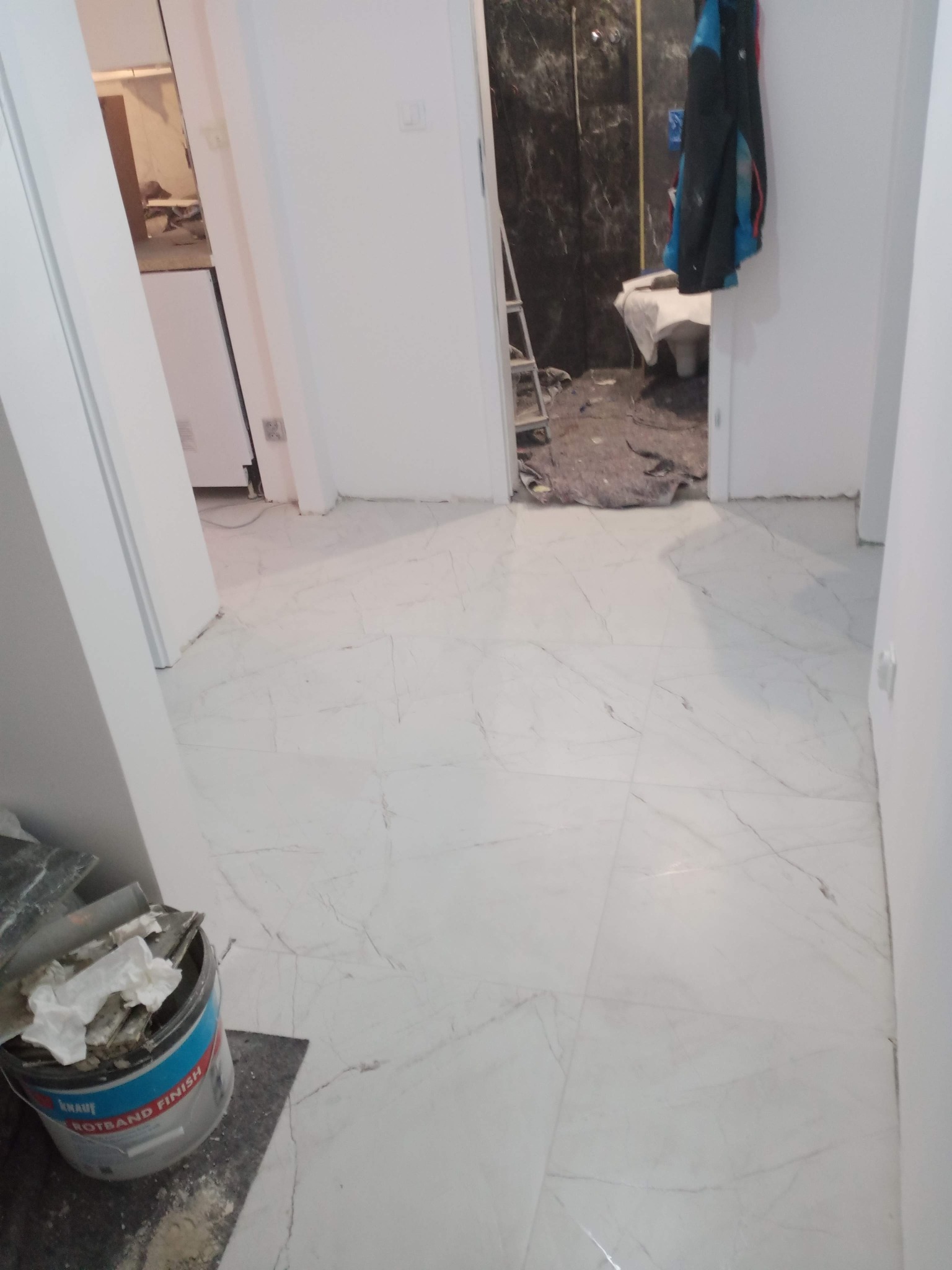 Wykończona podłoga z białego marmuru w nowym mieszkaniu. W tle widoczny remont łazienki, drabina i wiadro z zaprawą, co sugeruje świeżo zakończone prace wykończeniowe.