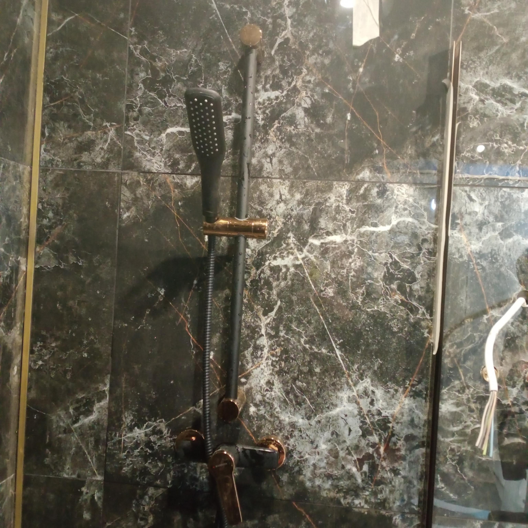 Czarny panel prysznicowy z złotymi akcentami na tle marmurowych płytek w kabinie. Widoczne przewody elektryczne i fragment szklanej ściany.