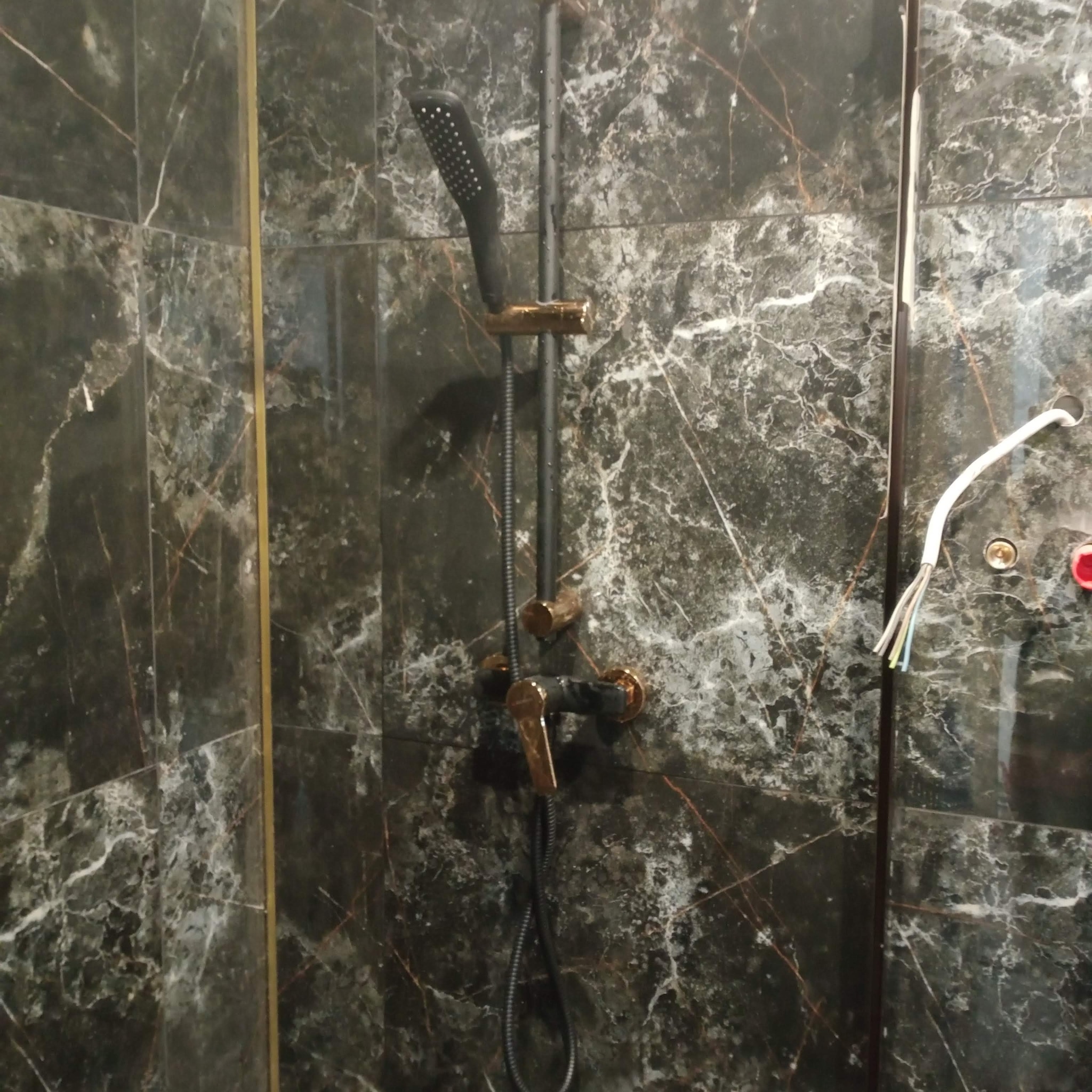 Czarno-złota armatura prysznicowa na tle płytek imitujących marmur. Widoczne przewody elektryczne, sugerujące etap montażu lub podłączenia.