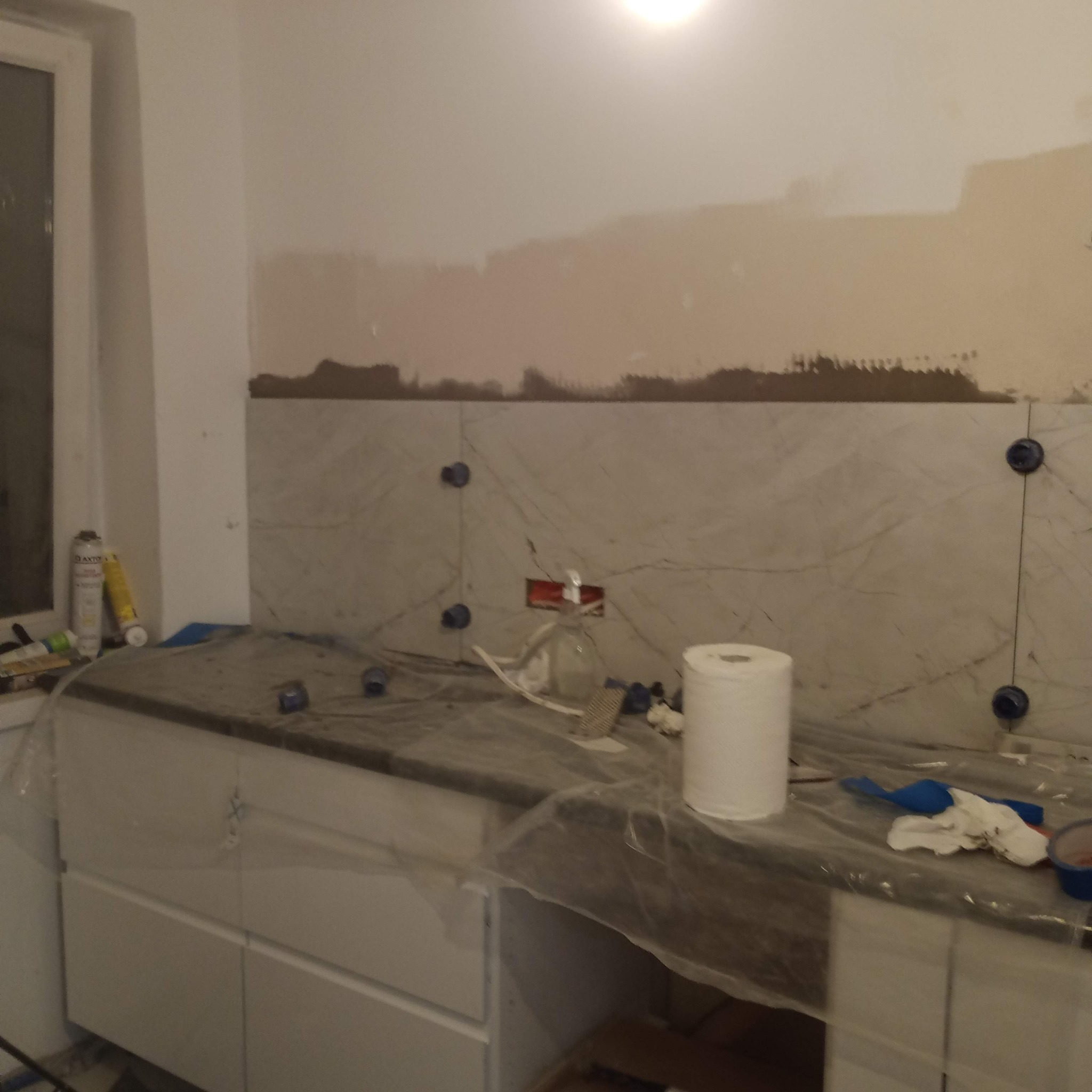 Układanie płytek marmurowych na ścianie w kuchni, widoczne krzyżyki dystansowe i częściowo przygotowana ściana. Blat zabezpieczony folią malarską.