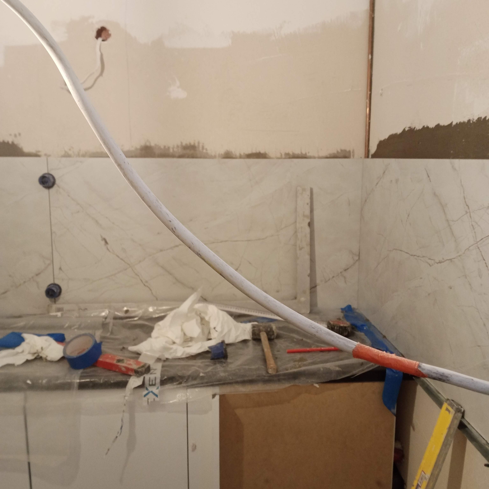 Remont kuchni: Ułożone płytki imitujące marmur na ścianie, widoczne narzędzia pracy, nieukończone podłączenie rur. Przygotowania do dalszych prac.