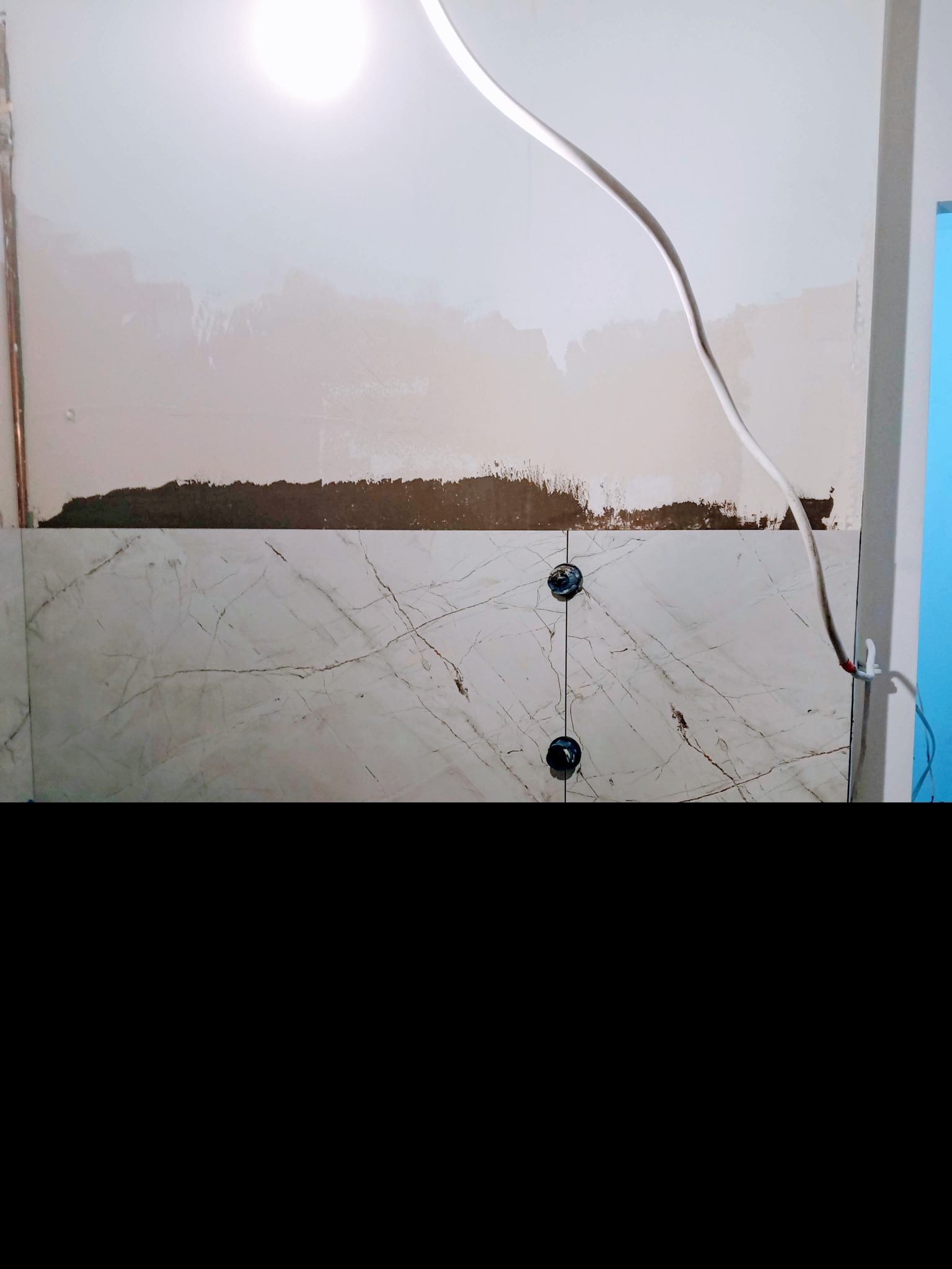 Montaż płytek imitujących marmur na ścianie, widoczne krzyżyki dystansowe i przewody elektryczne. Fragment ściany przygotowany do dalszych prac wykończeniowych.