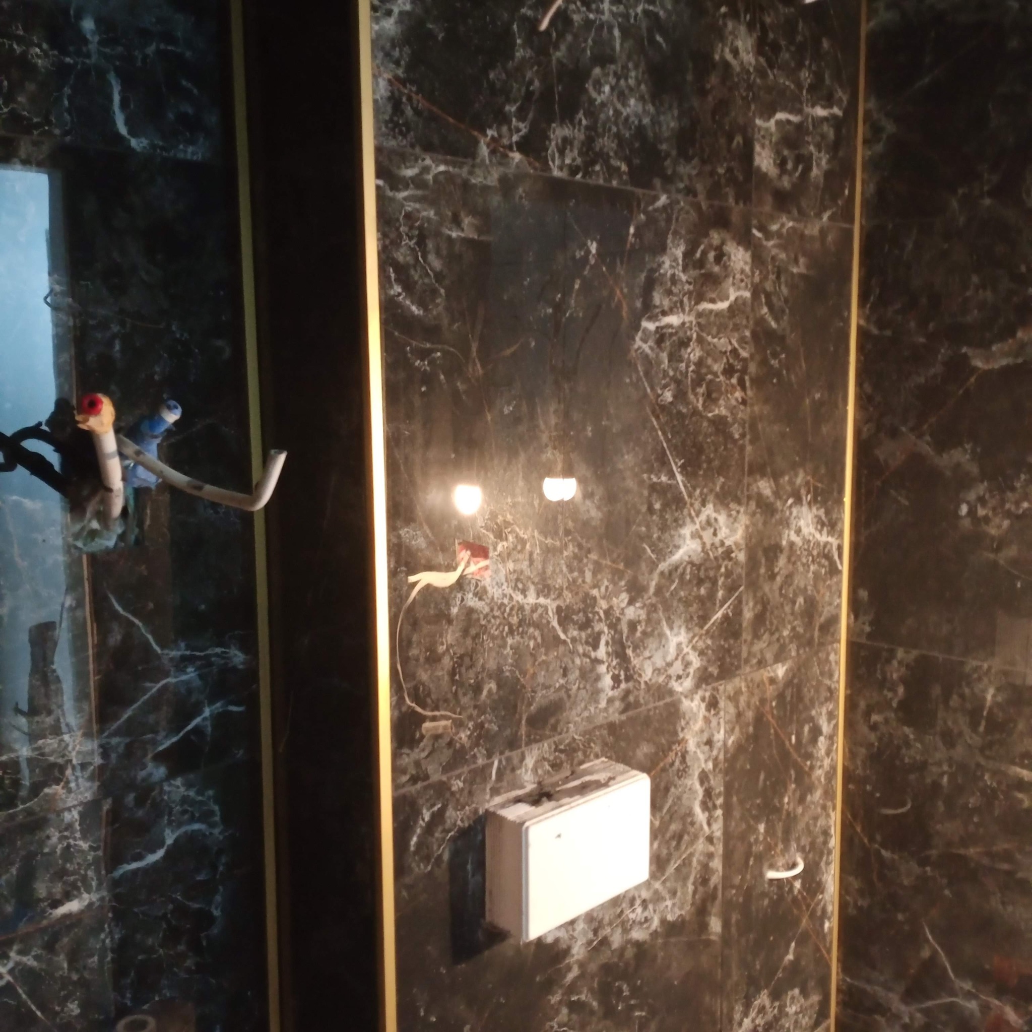 Częściowo wykończona łazienka z czarnymi marmurowymi płytkami i złotymi listwami. Widoczne wystające rury oraz instalacja elektryczna przed podłączeniem.