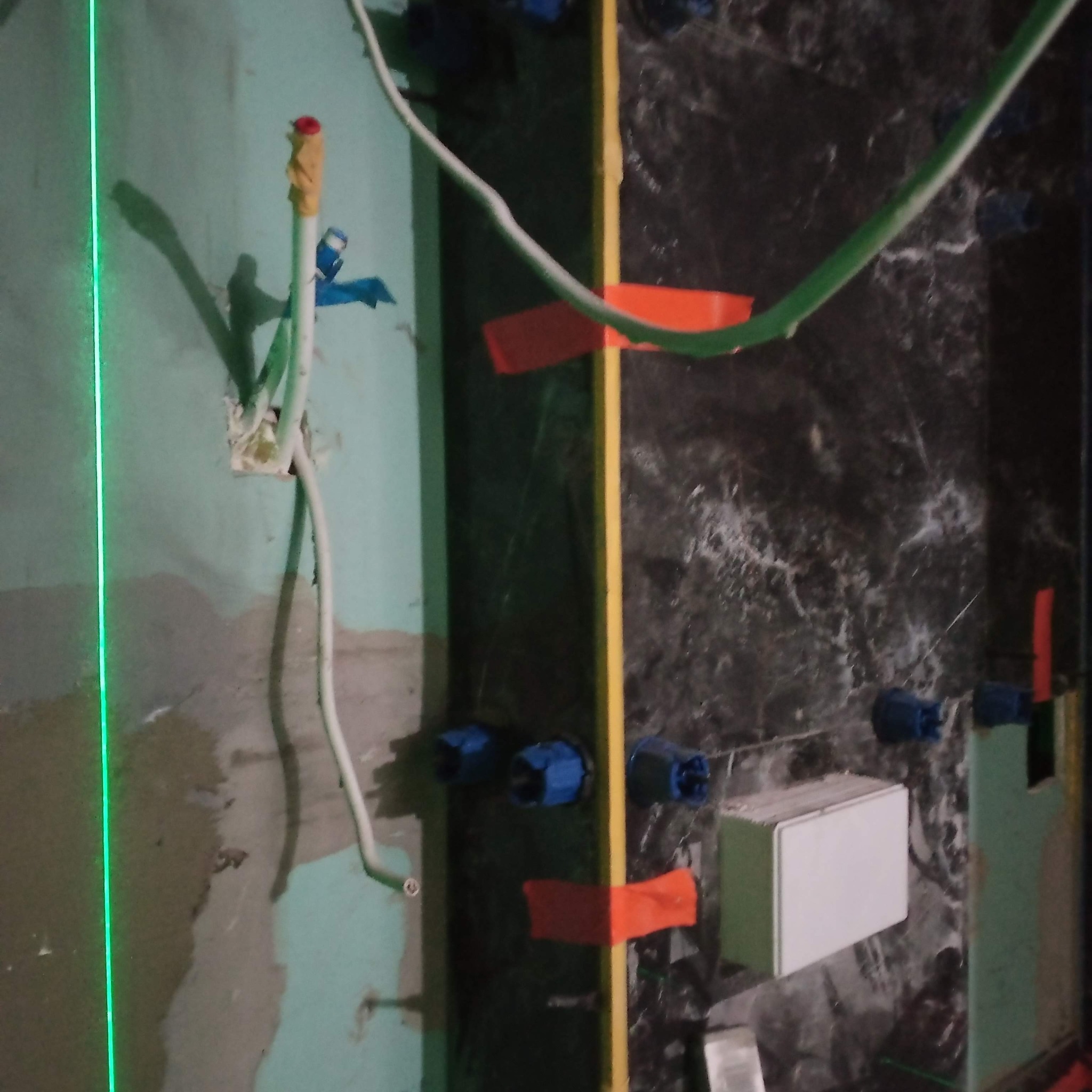 Instalacja wodna w trakcie montażu: rury, kable, poziomica laserowa. Widoczne elementy instalacji na tle ściany z płytkami imitującymi marmur.